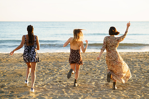 Full body back view of unrecognizable relaxed girlfriends running on sandy seashore during summer vacation at sundown