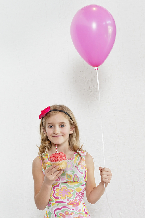 Portrait of happy girl holding cupcake and party balloon over colored background