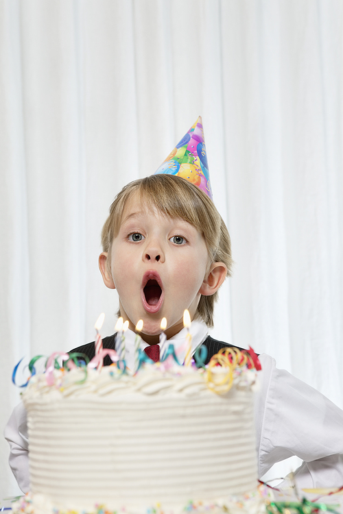 Portrait of young boy wearing party hat blowing candles on birthday cake
