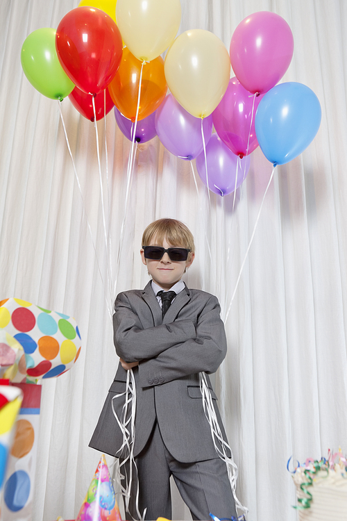 Young boy wearing sunglasses and standing with arms crossed holding party balloons