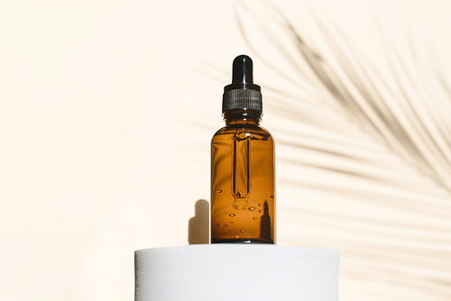 A dropper bottle of serum or liquid collagen on a beige background with a shadow from thin palm leaves. The concept of anti-aging cosmetics