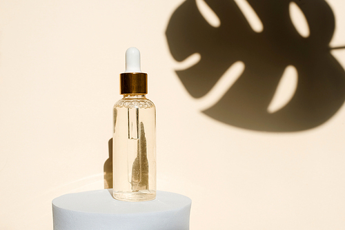 Essential oil, revitalizing and moisturizing serum on a podium on a beige background with the shadow of a tropical monstera leaf. Natural beauty product concept.