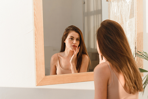 Attractive smiling young brunette looking in the mirror, touching and examining the face. Skin care concept