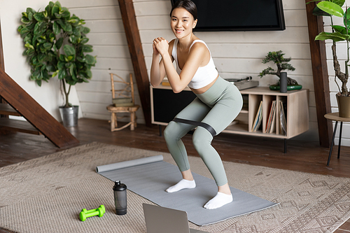 Cute asian fitness girl at home doing workout, squats with stretching elastic rope on legs, standing on floor mat, listening to online sport instructor on laptop video.