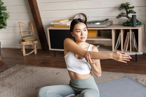 Asian fitness girl doing workout at home, sitting on floor mat in activewear, warm up and stretching arms, doing yoga in living room.