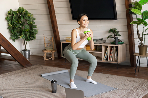 Fit and healthy asian woman doing squats at home, workout fitness training session with dumbbells and protein shaker in living room, watching sports video on laptop.