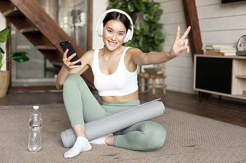 Happy fitness girl in headphones and activewear, holding mobile phone and showing v-sign, sitting near water bottle, workout at home.
