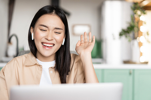 Happy asian woman laughing and smiling, talking on video call using laptop, waving hand and chuckling, wearing earbuds.