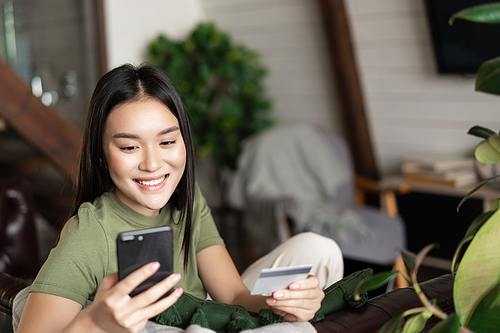 Smiling asian girl sitting at home with mobile phone and credit card, order in online shop, paying for delivery.