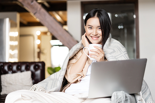 Smiling asian woman sitting at home wrapped in blanket, watching videos on laptop and drinking coffee or tea in mug.