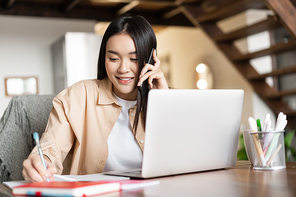 Smiling asian girl working from home, talking with client, answer phone call and taking notes in notebook, sitting near laptop.