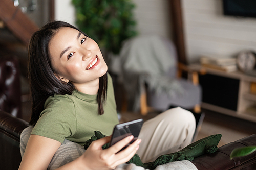 Dreamy smiling asian woman using mobile phone, sitting and relaxing at home.