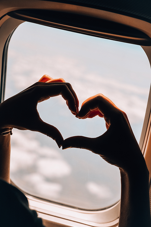 Female hand in heart shape Airplane flight. View from the window of the plane. Airplane, Aircraft. Vacation love travel concept Traveling by air. Airplane Window View Above the Clouds Amazing golden fluffy clouds moving softly on the sky aerial view.