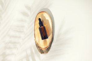 Amber glass bottle on wooden podium, white background with tropical leaves shadows, flat lay, top view. Ecological natural cosmetics concept