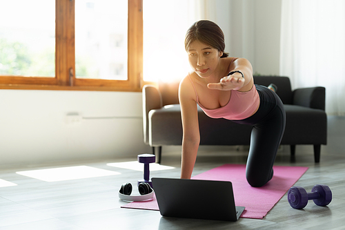 healthy sports asian woman in sportwear at home using laptop to share workout results in social media and doing abs exercises on fitness mat.