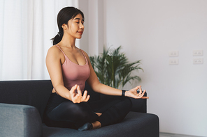 Young beautiful woman meditating on sofa at home interior sitting on yoga mat and smiling relaxed with closed eyes, Mindfulness meditation concept, Healthy Lifestyle.