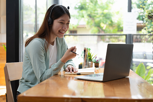 Smiling girl student wear wireless headphone study online with teacher, happy young woman learn language listen lecture watch webinar write notes look at laptop sit in cafe, distant education.