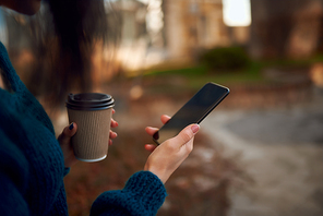 Cropped shot of young woman hands with a smartphone and disposable cup of hot drink on defocused urban background