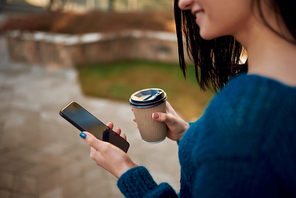 Close up of an unknown smiling female model looking on screen of a cellphone and drinking coffee outdoors
