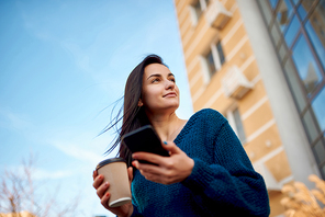 Young beautiful woman strolling along tall modern buildings and holding mobile phone and cup of coffee in hands