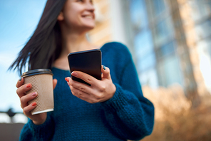Close up view of a black smartphone and disposable glass of coffee in hands of a blurry smiling young female