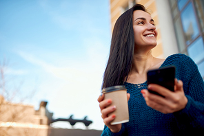Young woman with coffee and smartphone standing near the tall modern tower in a business district and enjoying cityscape and beautiful weather