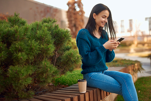 Smiling young woman sitting cross-legged on a wooden sidewalk of garden and comunicating with someone by telephone