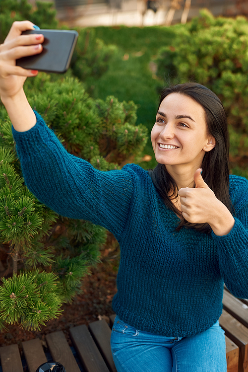 Nice-looking smiling young female doing photo of herself on her phone while showing thumb up gesture with green pines on background