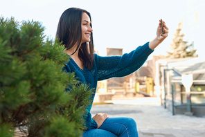 Side view of a smiling young female shooting selfie on her mobile phone near a nice thick dwarf pine