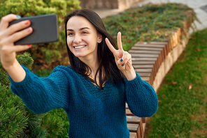 Trendy young woman taking positive selfie on a sidewalk of a green flowerbed, showing shiny smile and victory hand