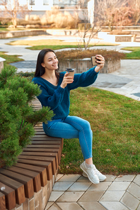 Smiling young woman in casual clothing sitting outdoors with a cup of coffee while making selfie