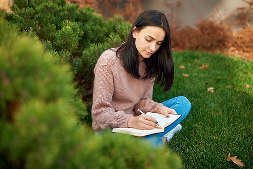 Pleasant young brunette with long hair is concentrated on writing on page of her notebook outside on an open lawn