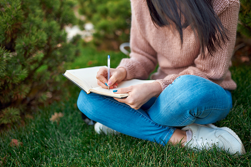 Close up of a young woman in jeans and sweater taking some notes in her paper planner while sitting on grass outdoors
