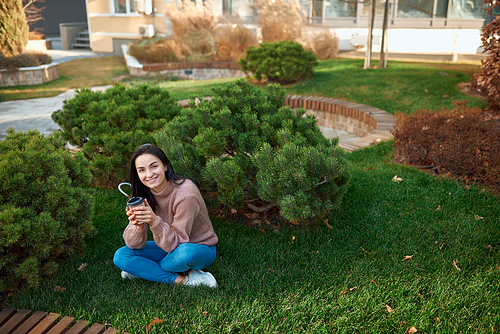 Cheerful young woman sitting cross-legged with cup of coffee in hands on fresh green grass in a front garden near the buildings
