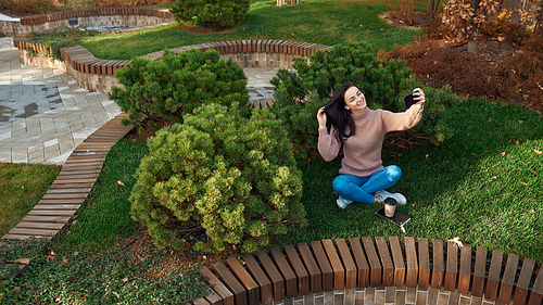 Charming young woman doing photo of herself on a cellphone while sitting cross-legged on grass and touching her long dark hair