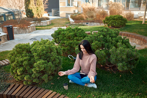High angle view of a pleasant young female relaxing in a meditative pose on a grass in cosy dooryard in front of modern edifices