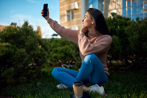 Beautiful young female in jeans and sweater making photo portrait of herself on a mobile phone outdoors