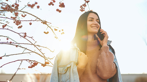 Charming young lady standing outdoors in sunlight and having pleasant conversation by cellphone