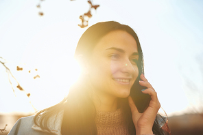 Close up portrait of a smiling young female holding her smartphone near ear outdoors in a warm autumn evening