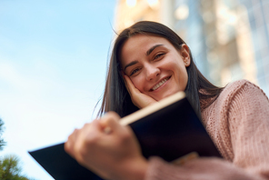 Beautiful young female looking over the pages of book and holding in hand with smile on blurred background