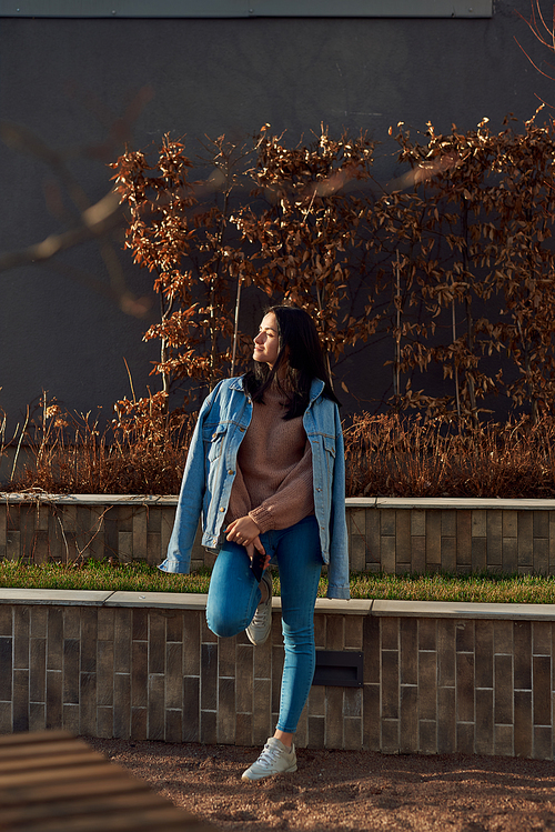 Beautiful young lady in denim clothing standing relaxedly in an autumn square, basking in warm sunlight