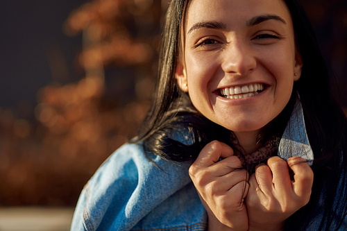 Close up of a smiling young female brunette wrapping herself up in a jeans jacket on defocused background outdoors