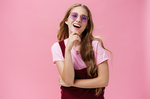 Self-confident good-looking pretty young lady with wavy hair floating on air raising head up having high self-esteem touching chin laughing and smiling wearing trendy sunglasses and dungarees. Body language and emotions concept