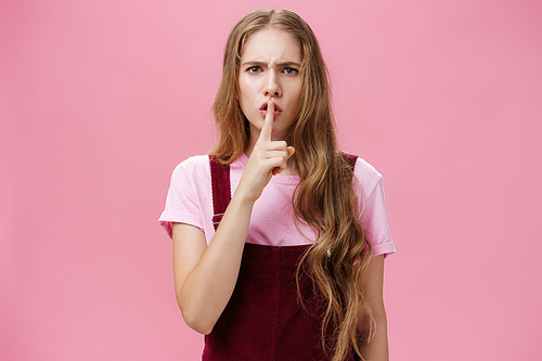 Girl displeased by loud guys speaking in library shushing at camera with dissatisfied expression making shh gesture with index finger over mouth wearing trendy corduroy overalls against pink wall. Body language concept