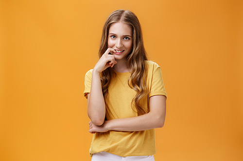 Creative tricky young woman with natural wavy long hair in yellow t-shirt looking from under forehead with intention and lust in expression biting finger, smiling at camera over orange wall.