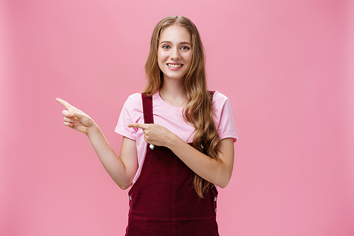 Polite and charismatic pretty young girl with long fair hair in corduroy trendy overalls pointing left as if inviting or showing something interesting smiling friendly at camera over pink background.