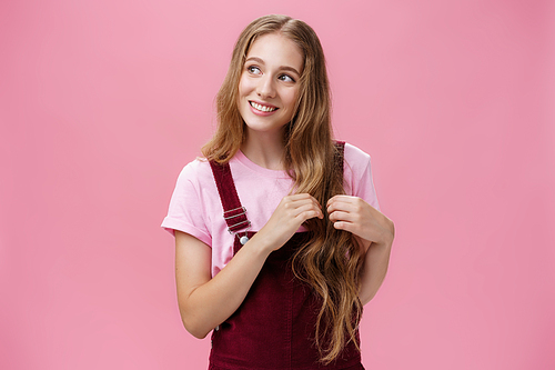 Silly kind and friendly-looking charming girl daydreaming picking strands of hair and looking left with nice cute smile imaging, picturing lovely scene posing flirty and happy over pink background.