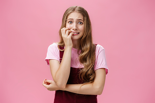 Woman panicking and overreacting feeling nervous biting fingernails and staring afraid at camera trembling from fear over pink background posing in cute dungarees with natural wavy hairstyle.