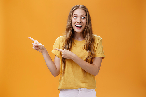 Portrait of wondered and amazed young excited girl with scar on arm and wavy hair open mouth in enthusiasm and joy pointing left amused and questioned eager to watch show over orange wall. Advertising, emotions and people concept