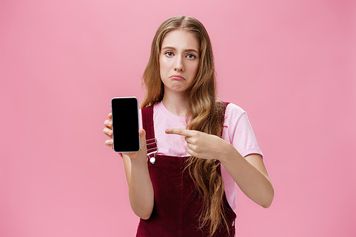 Sad gloomy young female with cute wavy natural hairstyle showing smartphone screen pointing at gadget with index finger making upset face frowning displeased feeling regret after buying broken phone.